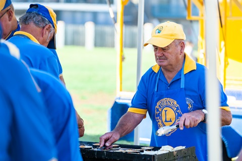 Lions Club member, man, wearing all blue and yellow (Lion's Club branding) and cooking a bbq holding tongs