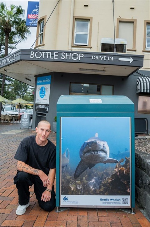 photograph of a shark printed onto the side of a bin in Kiama main street. Photograph with the artist, a young man, dressed in all black, kneeling beside bin.